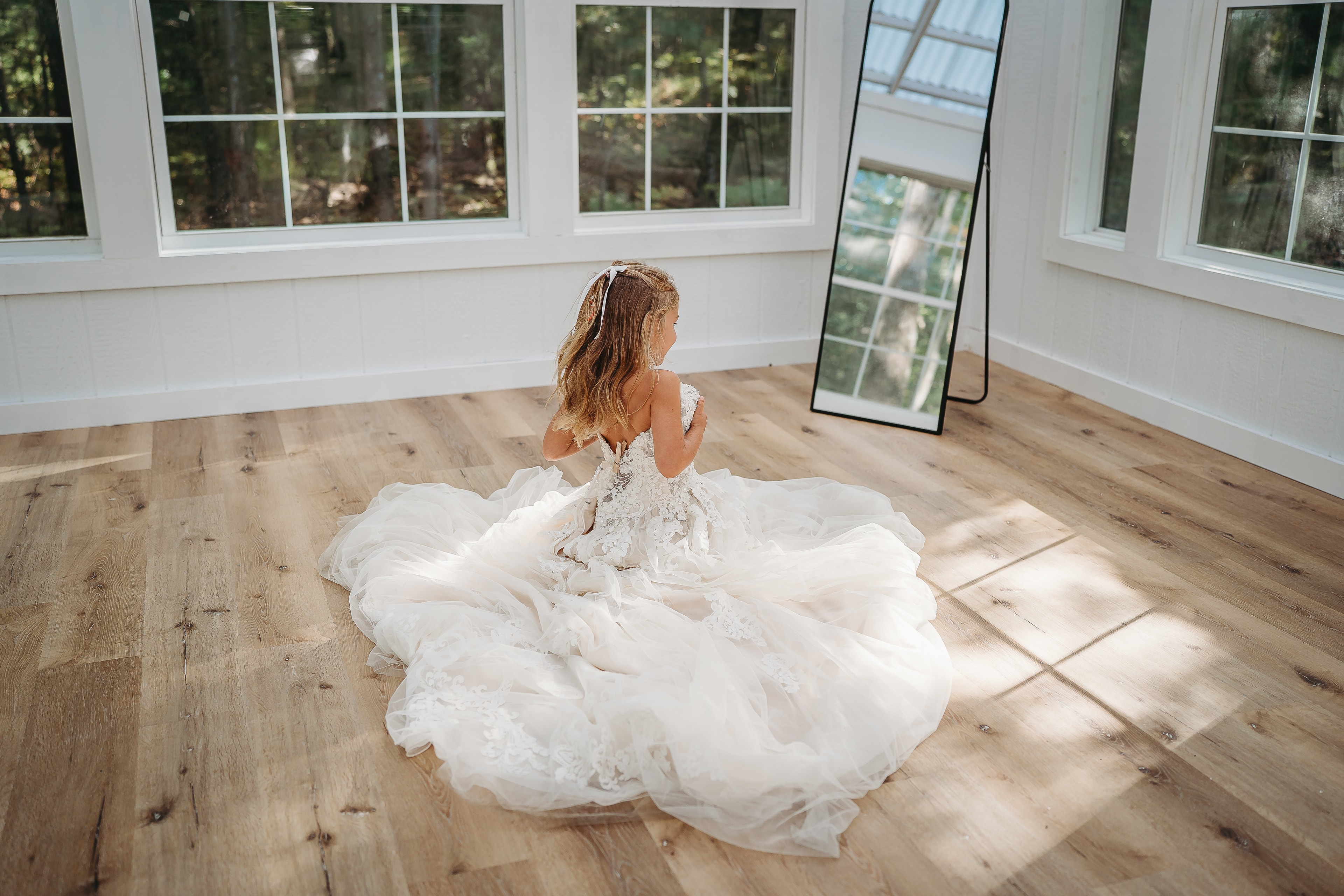 Little girl in an oversized gown sitting on the studio floor near a tall mirror